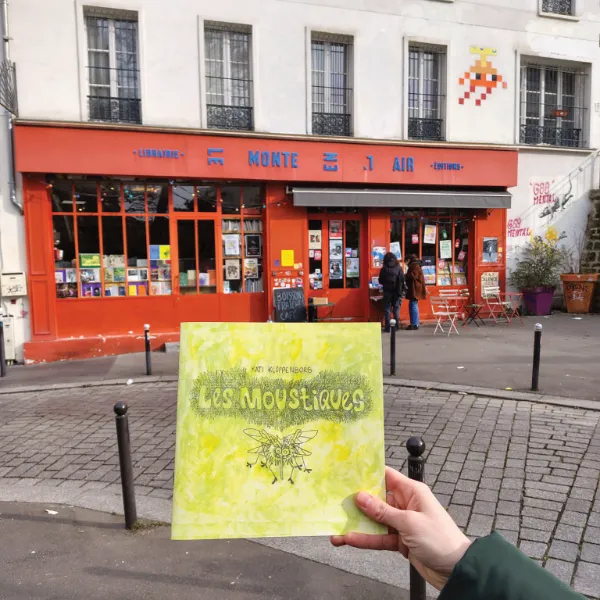 In the foreground: a hand holding a print version of the comic. In the background :  a bookshop